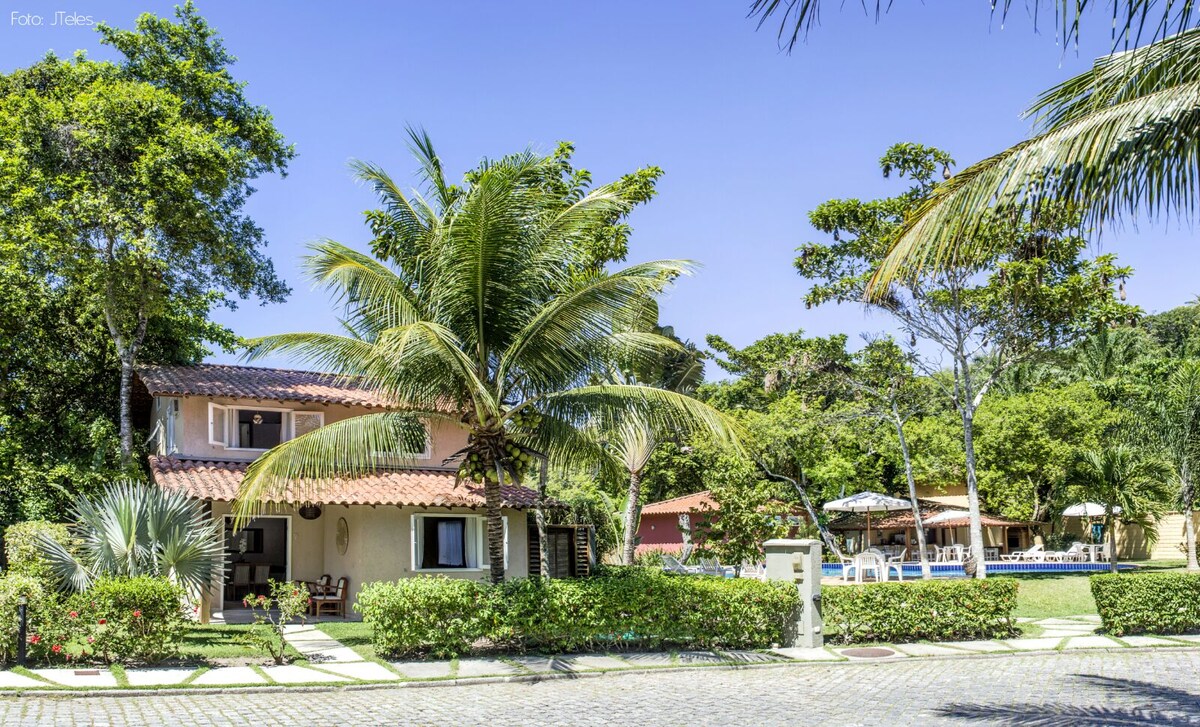 A two-story house is showcased, surrounded by lush palm trees and greenery. The cobblestone path leads to the entrance, while an inviting pool area is visible in the distance, along with shaded seating under umbrellas in the expansive garden area.