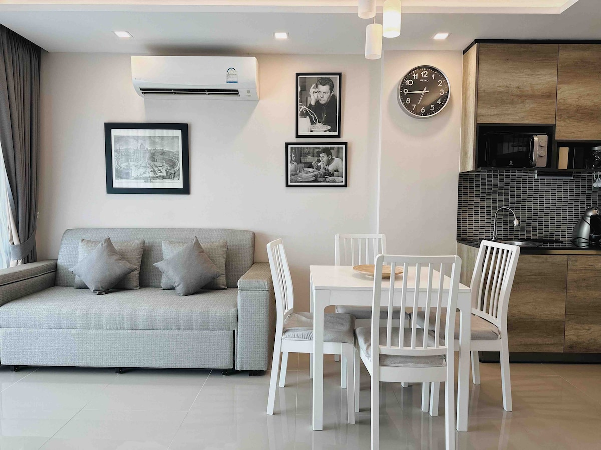 A modern living space featuring a light grey sofa adorned with decorative cushions. A dining table with four white chairs is positioned nearby. Artwork hangs on the wall, alongside a clock. The kitchen area showcases cabinetry and appliances, blending functionality and style.