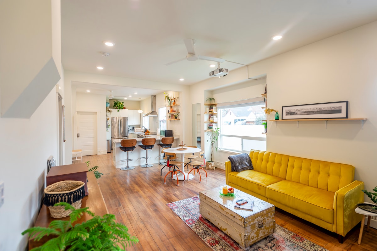 An open living area features a bright yellow couch, a wooden coffee table, and a rug that adds texture. The kitchen is visible in the background with modern appliances and bar seating. Natural light floods through large windows, creating an inviting atmosphere.