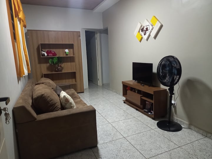 Dnevni boravak sa zajedničkom sofom, policom pored kreveta, stalkom za multimediju s televizorom i podnim ventilatorom.