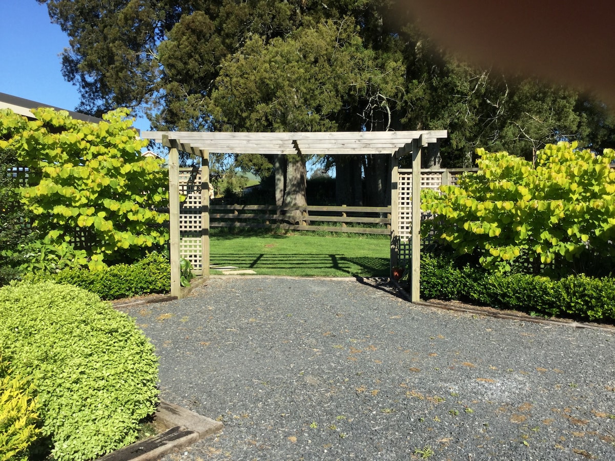 A wooden archway is framed by lush greenery, leading to an open grassy space. The path is lined with neatly arranged gravel, complemented by well-maintained hedges. Shade is provided by nearby trees, creating a peaceful entry point to the property.