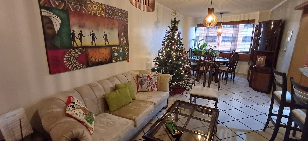 A comfortable living area features a beige sofa adorned with decorative cushions. A glass coffee table is centered on the tiled floor, and a vibrant artwork hangs on the wall. A Christmas tree is decorated and plants are arranged beside a window allowing natural light.