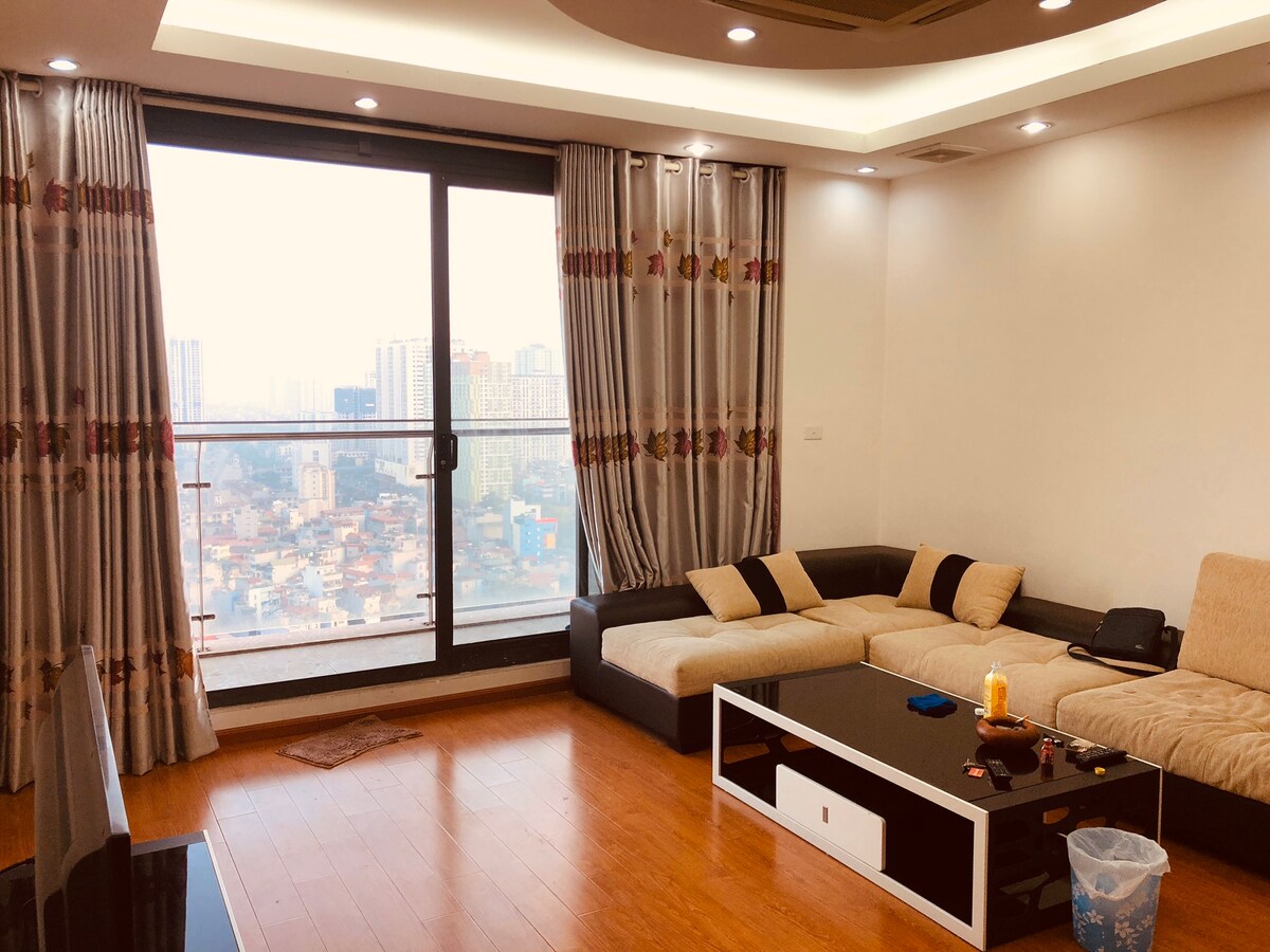 A spacious living room is featured, with large windows offering expansive city views. A modern sectional couch is positioned in front of a sleek coffee table, while warm wooden flooring complements the soft colors of the furnishings. Natural light fills the area, creating an airy ambiance.