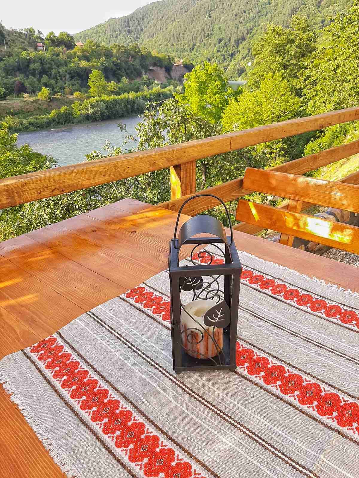 A wooden balcony table is set with a decorative lantern featuring intricate designs. A candle is placed inside the lantern, and a colorful woven table runner adds texture. The natural scenery of lush hills and a river is visible in the background.