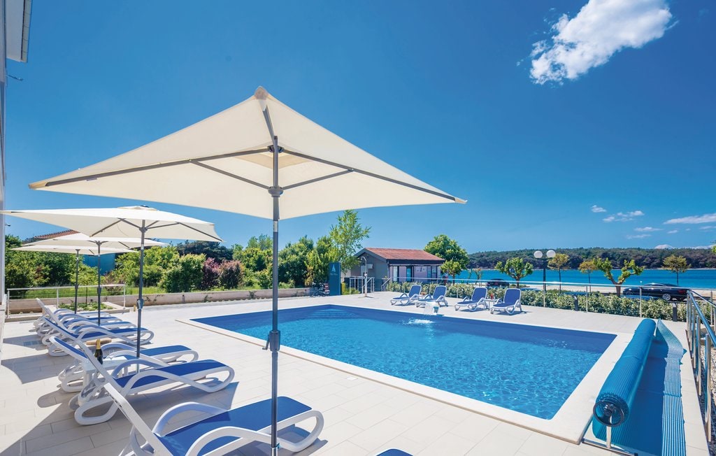 An outdoor swimming pool features a clear blue surface, surrounded by multiple loungers beneath large umbrellas. Lush greenery and distant views of the coast are visible in the background, complemented by a bright blue sky.