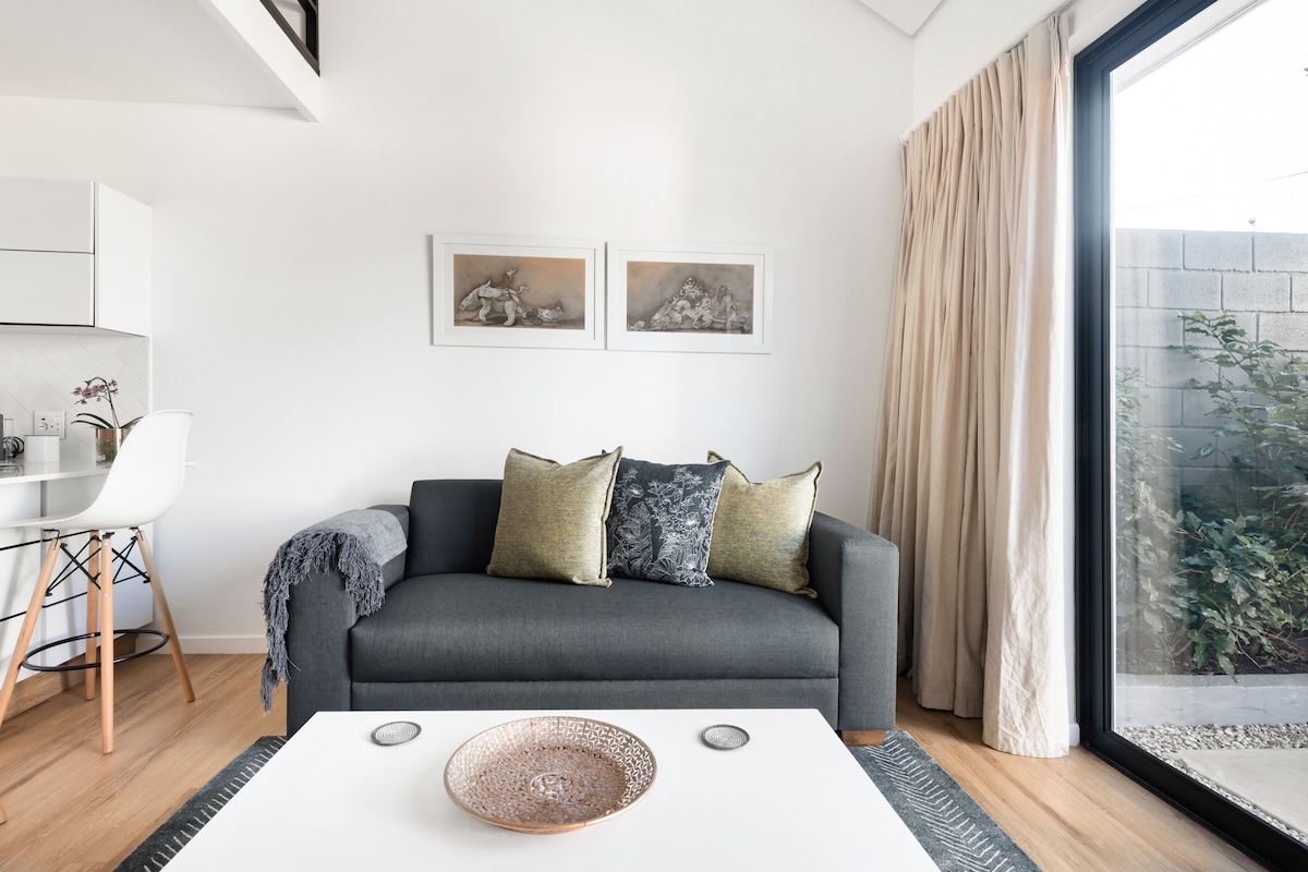 A modern living area features a dark grey sofa adorned with accent pillows in shades of green and blue. A light coffee table sits in front, with a woven bowl placed centrally. Large windows allow natural light to fill the space, framed by light-colored curtains.