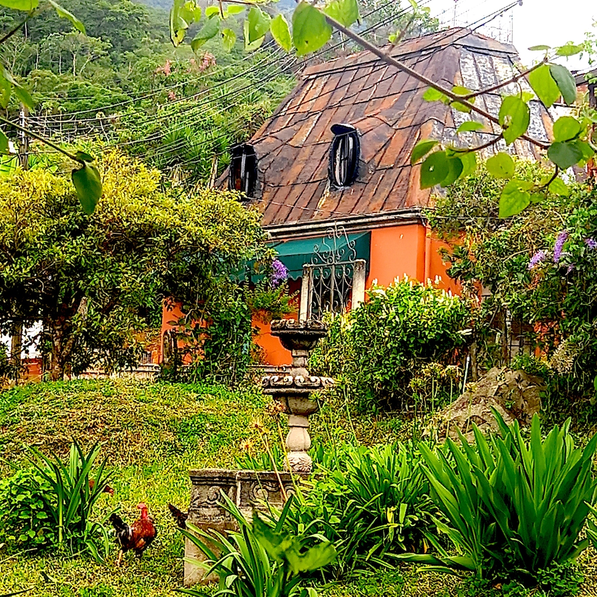 A charming casita is nestled among lush greenery, featuring a rustic, weathered roof and vibrant orange walls. A decorative stone fountain is surrounded by flowering plants, with chickens roaming nearby. The backdrop showcases the serene mountains, enhancing the peaceful ambiance of the outdoor space.