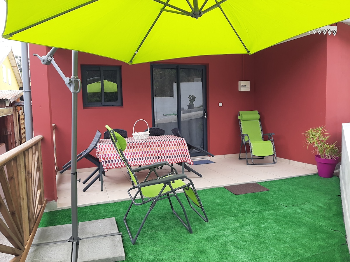 A covered terrace with a bright green umbrella provides shade over outdoor seating. A table is set with a red and white patterned tablecloth, flanked by two reclining chairs. Lush artificial grass adds a vibrant touch to the outdoor space, creating a relaxing atmosphere.