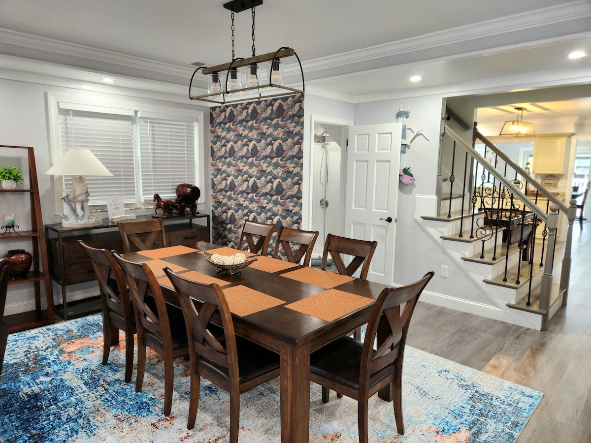 A spacious dining area features a large wooden table, comfortably seating eight. Colorful placemats are arranged on the table, complemented by a decorative centerpiece. Bright lighting illuminates the room, and a patterned wallpaper adds visual interest to the backdrop.