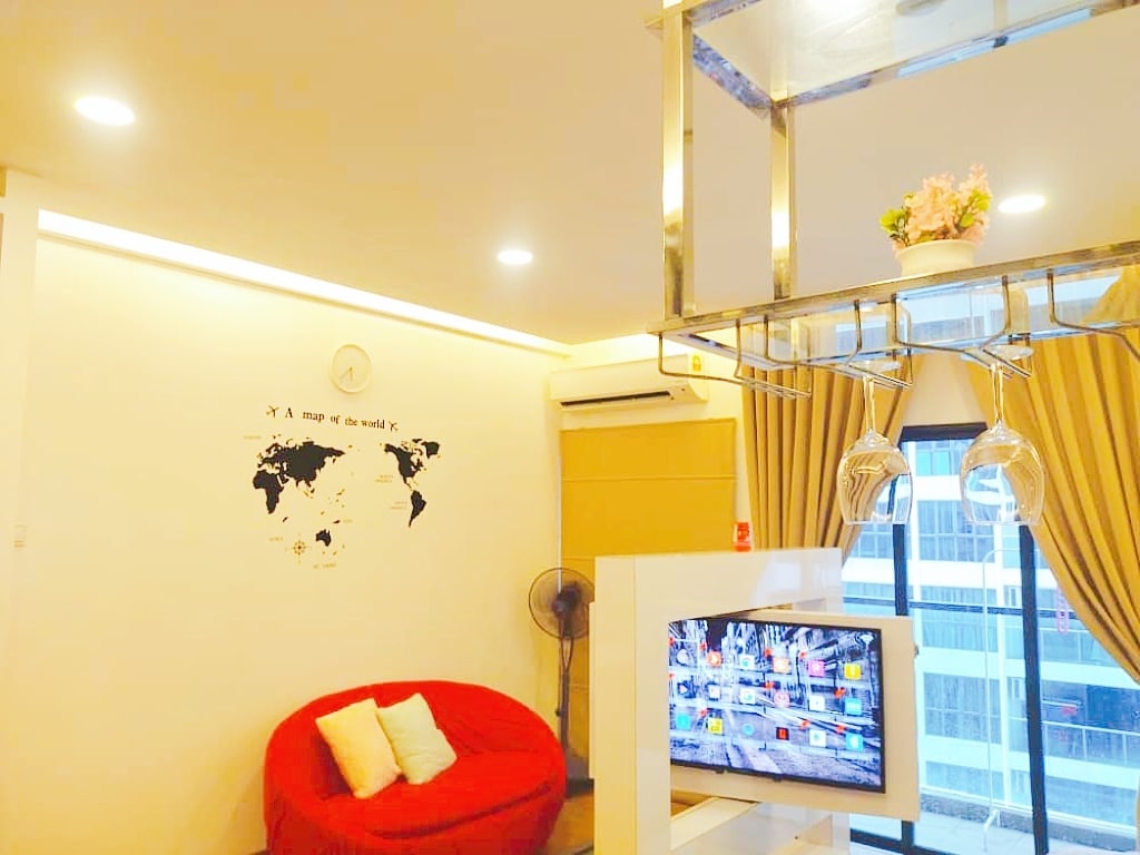 A cozy seating area is highlighted by a bright red chair, positioned near a wall featuring a world map design. A television with various apps is placed on a standing unit, while light from large windows complements the warm ambiance created by soft lighting.