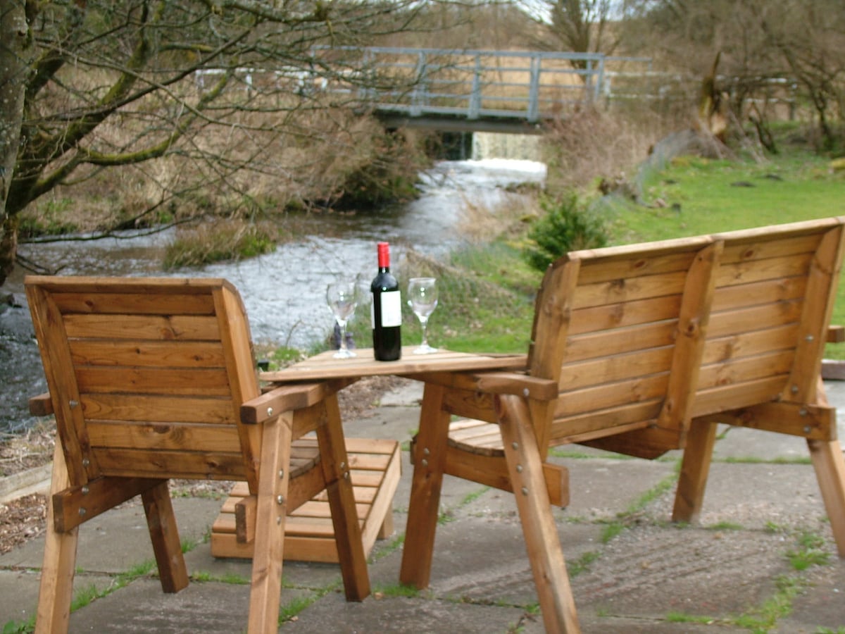 Two wooden chairs are positioned at a small table near a flowing river. A bottle of wine and two glasses rest on the table, while a scenic view of an adjoining bridge and lush greenery can be seen in the background.