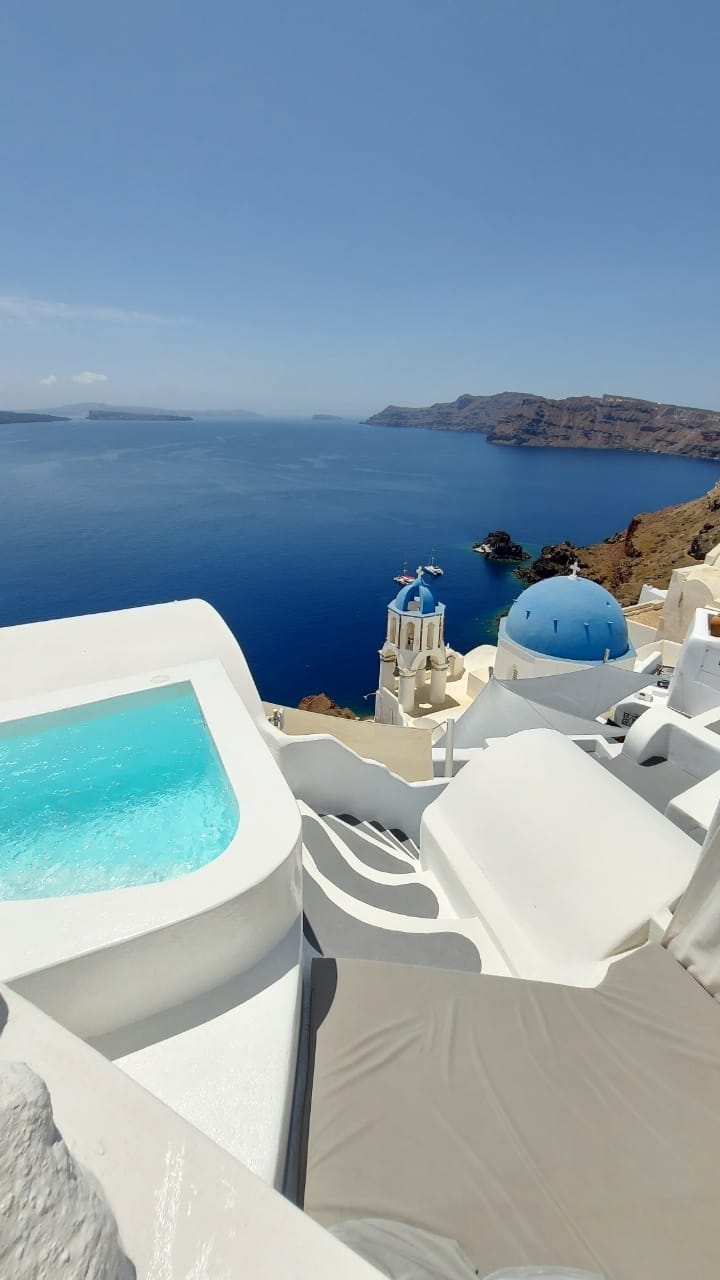A sparkling private heated pool is prominently situated on a terrace, with panoramic views of the deep blue sea and coastline. Nearby, a whitewashed dome of a church and a vibrant blue dome contrast against the clear sky, enhancing the serene surroundings.