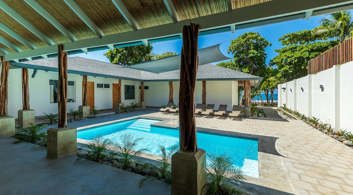 A spacious outdoor area is presented, featuring a sparkling pool surrounded by lounge chairs. Natural light fills the space, with greenery visible in the background. The design incorporates wood elements and a shaded seating area, creating an inviting environment for relaxation.