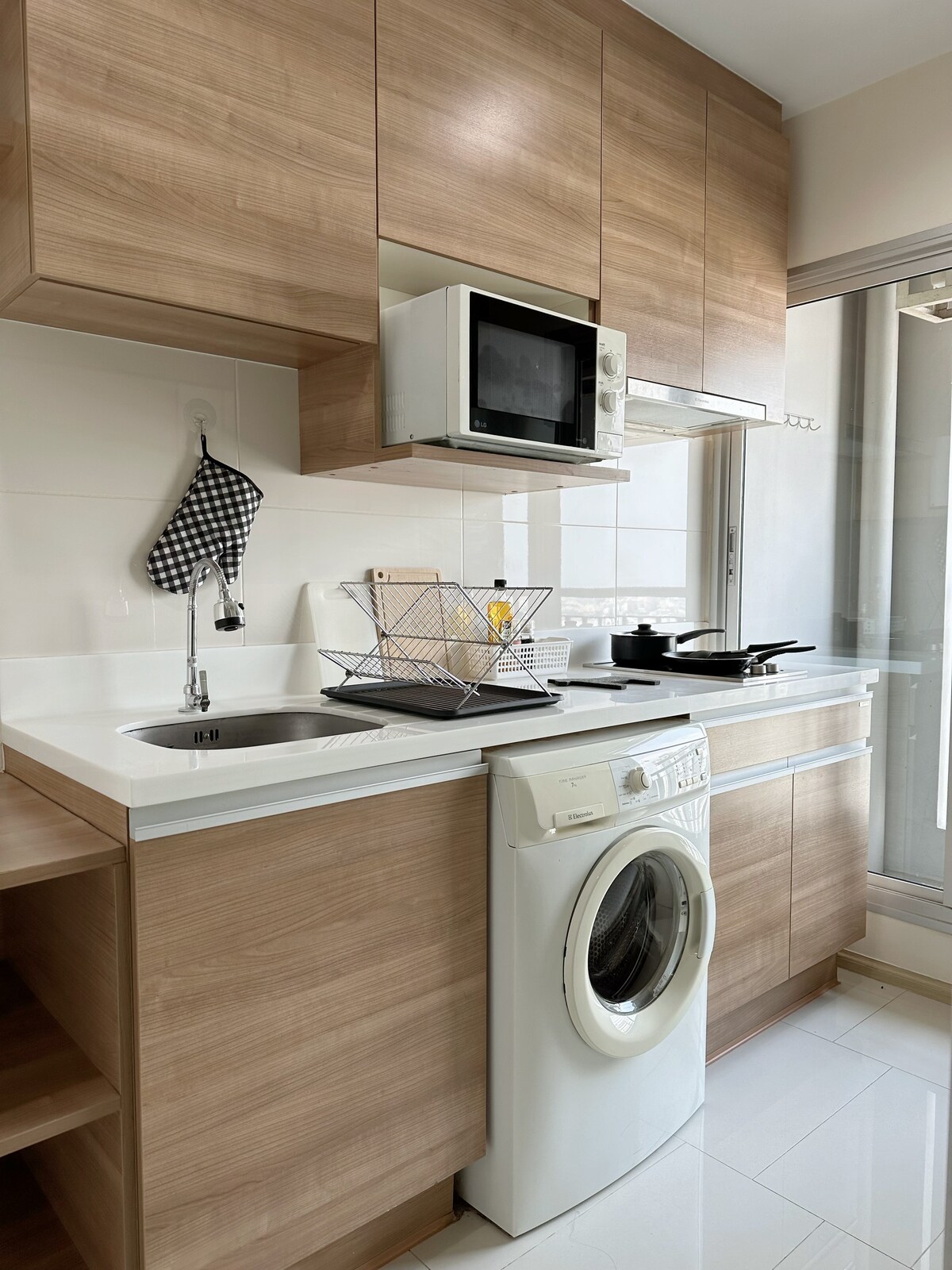 The kitchen features wooden cabinetry and a modern design. An induction stove, microwave, and refrigerator are visible. A sink is located beneath a window, with kitchen utensils organized in a drying rack. A washing machine is placed at the base, providing convenience for laundry.