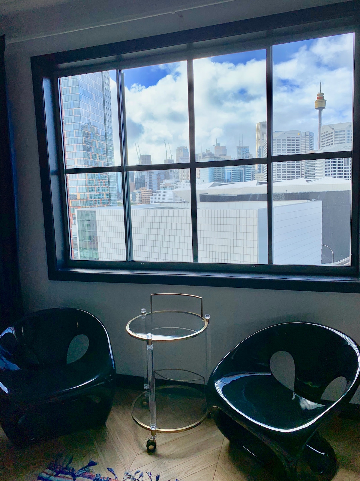 Two modern black chairs are positioned beside a gold accent table, creating a cozy seating area. A large window offers a clear view of the city skyline, featuring prominent buildings and the Sydney Tower, with a bright sky filled with clouds in the background.