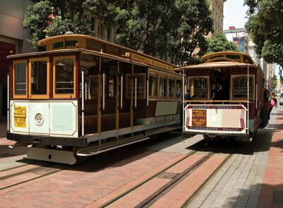 Two classic cable cars are parked side by side on a city street, with their distinctive wooden exteriors featuring vibrant colors and detailed signage. The surrounding area includes tree-lined sidewalks, contributing to the urban ambiance.