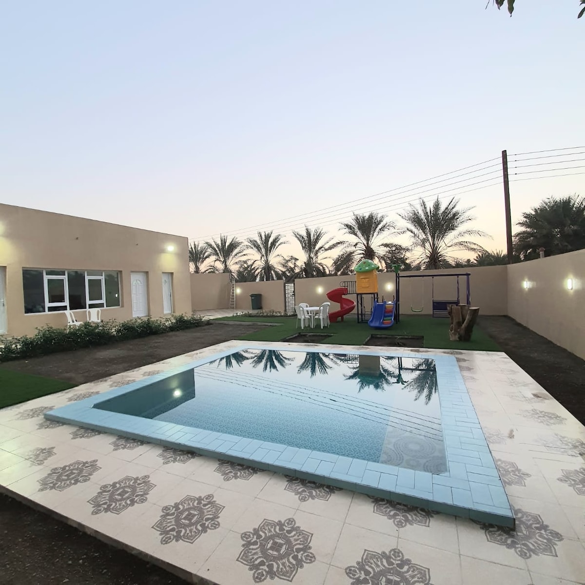An outdoor area showcases a pool surrounded by well-maintained grass. Nearby, a playground features a slide and swings. The walls of the house are painted in neutral tones, with palm trees visible in the background. Evening light enhances the tranquil setting.