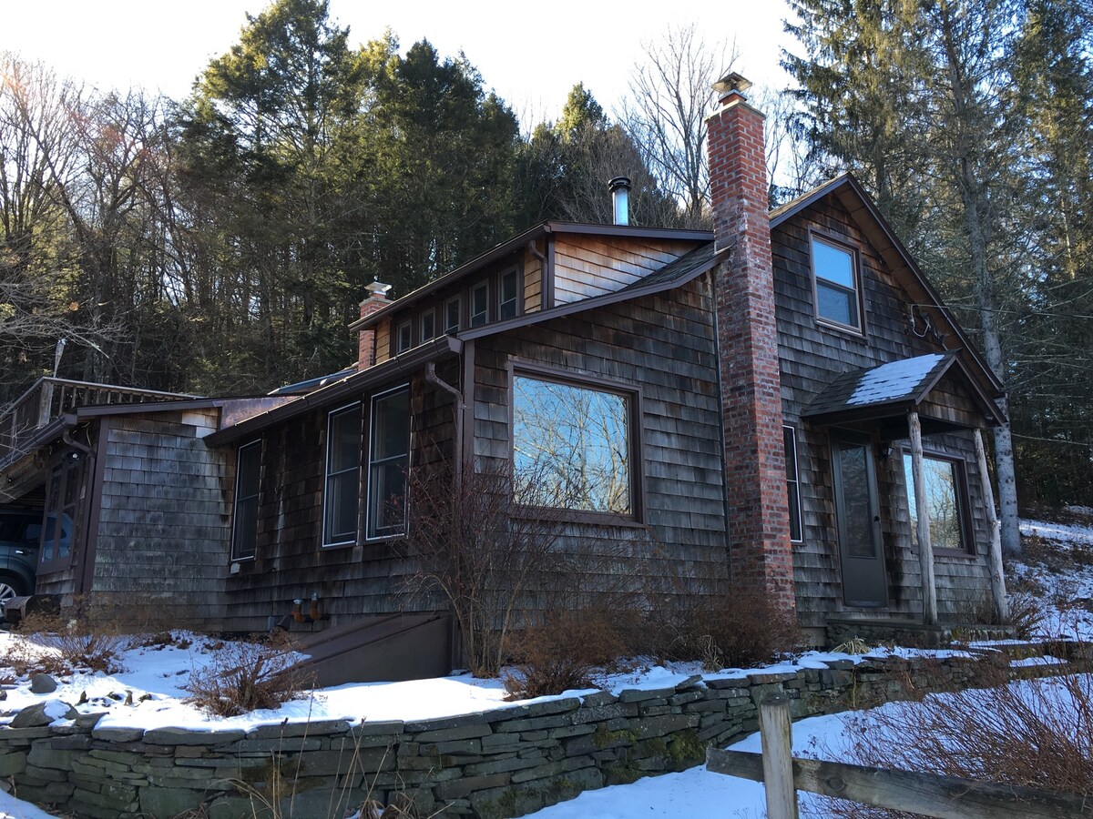 A charming house stands amidst a snowy landscape, featuring a combination of wooden shingles and stone accents. Large windows allow natural light to enter, while a chimney rises above the roof. Surrounding trees are visible in the background, providing a serene mountain setting.