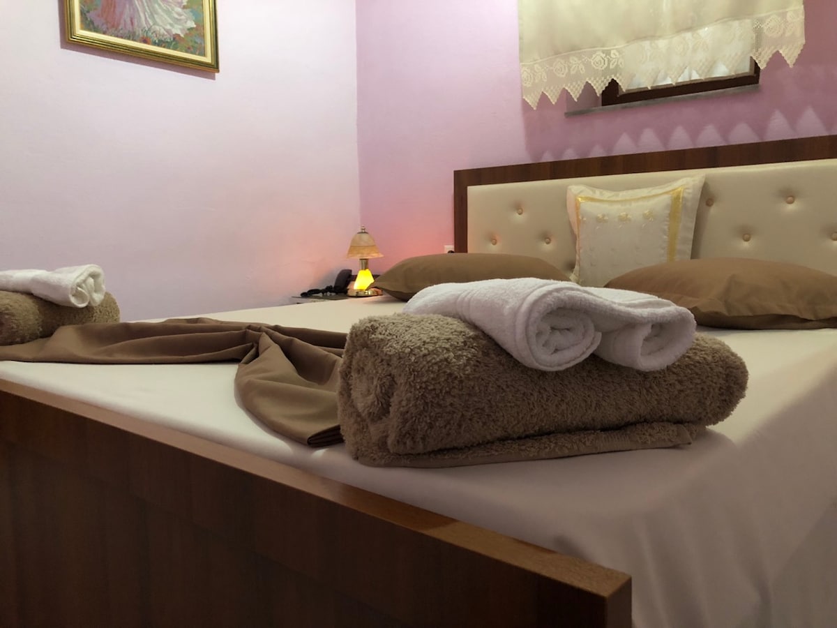 A cozy sleeping area features a plush bed with neutral-toned bedding and decorative pillows. Rolled towels are neatly arranged on a soft, textured throw. Soft lighting creates a warm ambiance, while a pastel wall adds a calming touch to the space.