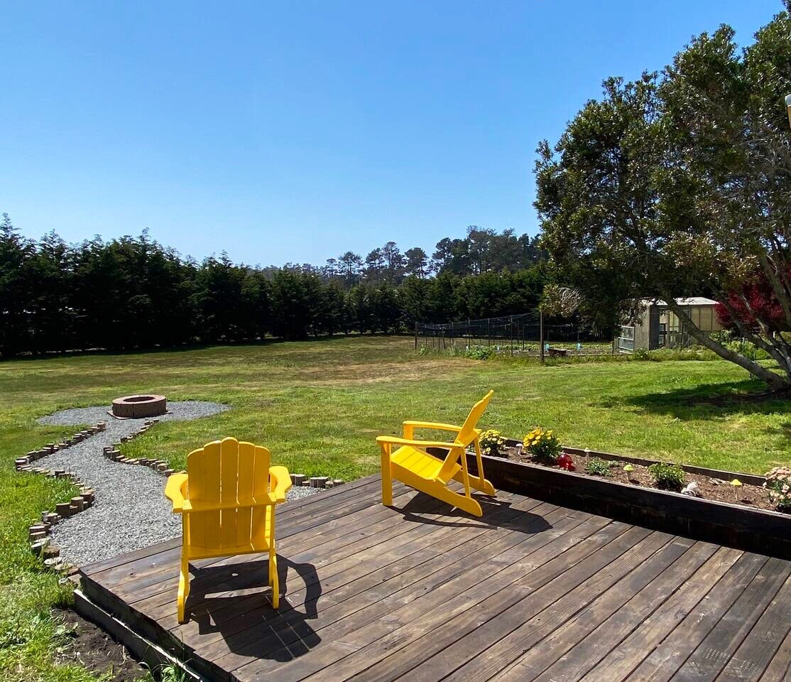 A wooden deck features two yellow chairs facing an expansive green yard. A winding gravel pathway leads towards a fire pit area. Mature trees border the property, providing a natural backdrop under a clear blue sky.