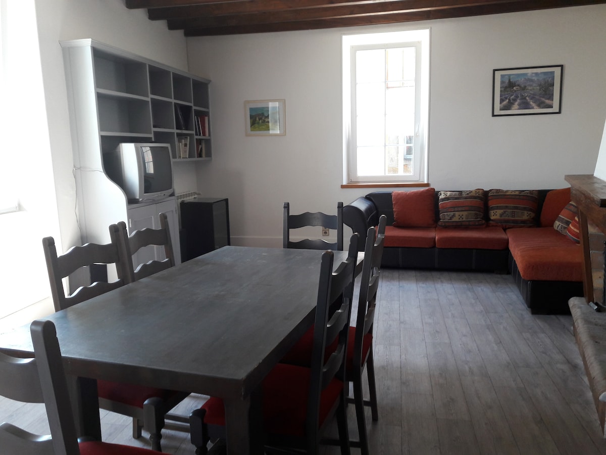 A welcoming living area features a large dining table surrounded by chairs and a comfortable sectional sofa. Natural light fills the space through tall windows. Shelving units and a television are situated along the walls, creating a functional and inviting atmosphere for relaxation.