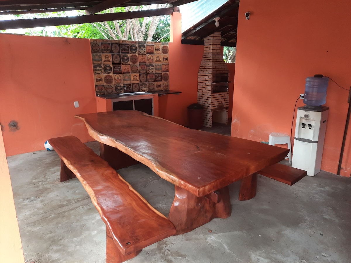 A large wooden dining table with a natural edge is complemented by a matching bench. The table is set in a semi-open area with orange walls, featuring a rustic brick grill and a wall adorned with wooden plaques in the background.