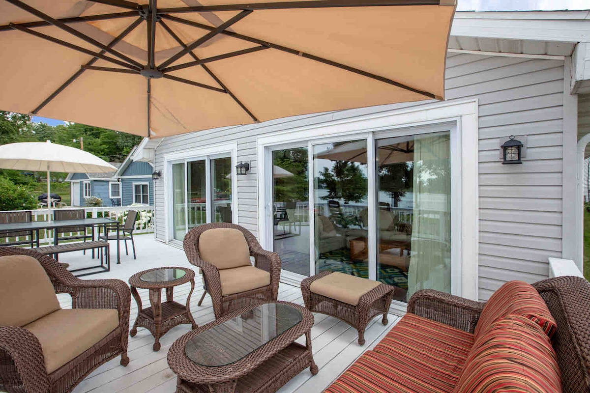 A spacious outdoor deck is presented, featuring comfortable seating arrangements with a large umbrella for shade. A sectional sofa and multiple armchairs surround a glass-top table. Large sliding glass doors lead into the interior, enhancing the connection to the serene lake setting.