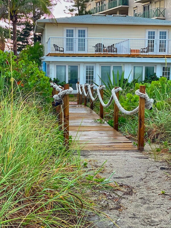 A wooden pathway is lined with lush green vegetation, leading towards a light-colored building. The pathway features decorative ropes along the sides. The area exudes a serene coastal vibe, with the oceanfront visible beyond the property.