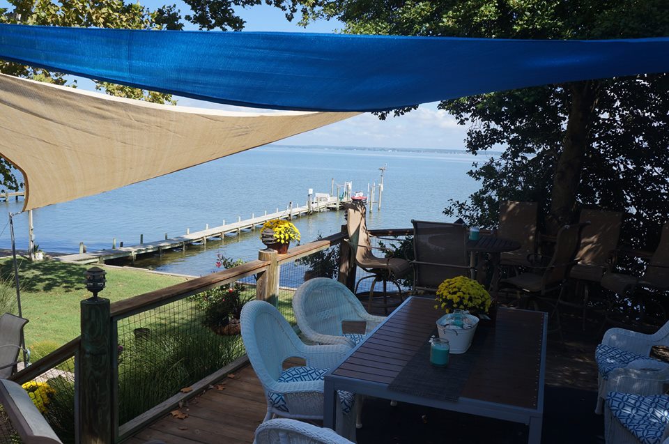 A shaded outdoor seating area features comfortable chairs and a table, overlooking the Rappahannock River. Brightly colored flowers adorn the space, while a long pier extends into the water, inviting relaxation and scenic views.