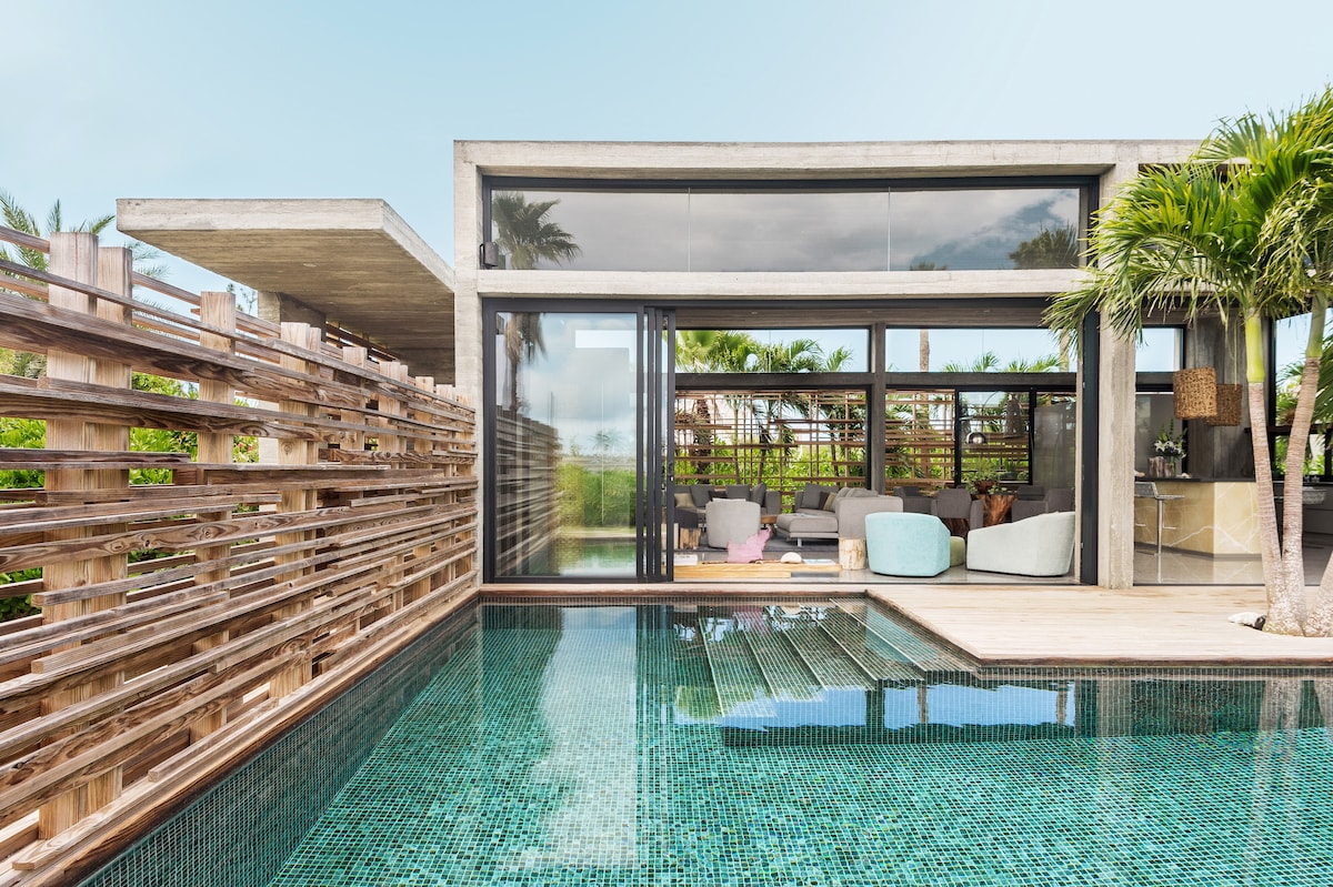 A modern villa features large glass walls, providing an open view of the spacious interior from the outdoor deck. A reflecting pool is situated in front, with tropical plants surrounding the area. Colorful lounge seating creates a relaxed atmosphere near the pool.