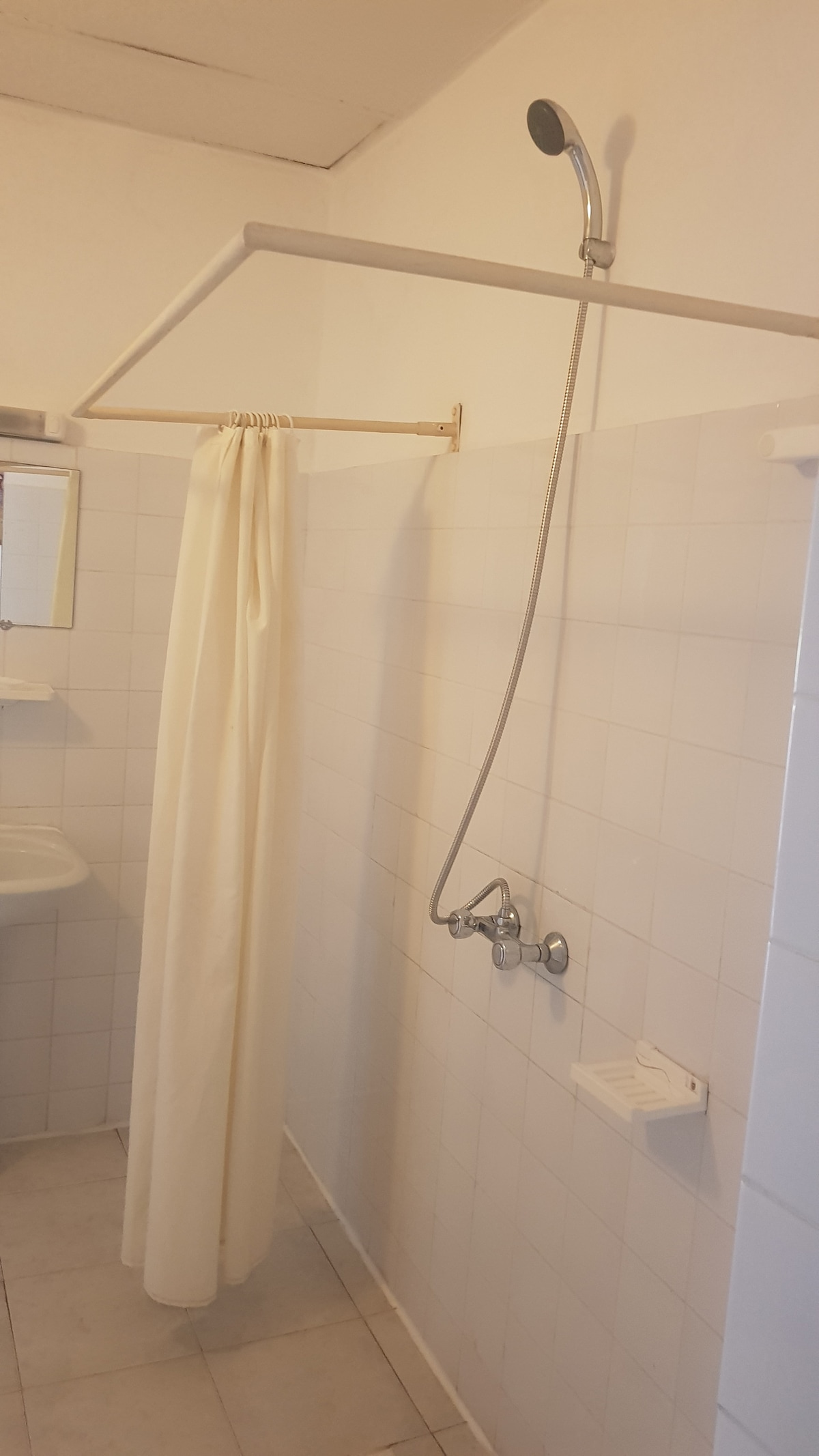 A clean bathroom is presented, featuring a mounted showerhead above a simple shower curtain. A small sink is visible to the left, accompanied by a mirror above it. The tiled walls and floor are in neutral colors, creating a calming environment.
