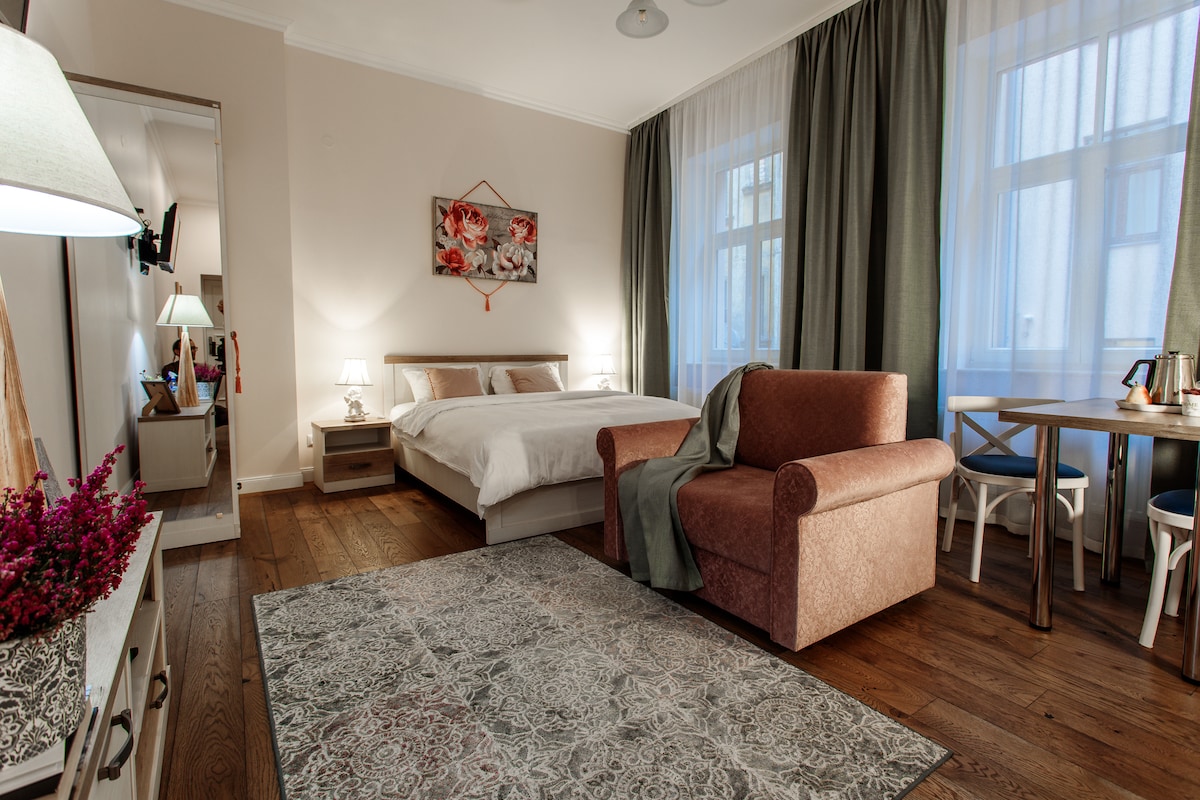 A spacious studio room features a kingsize bed and a comfortable chair. A dining table is present, and natural light filters through sheer curtains. A decorative rug adds warmth to the wooden floor, while a mirrored cabinet enhances the sense of space.