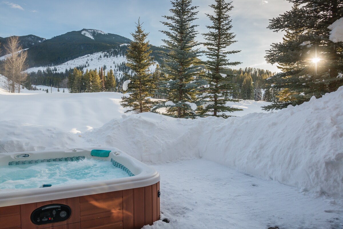A private hot tub is situated on a snow-enclosed deck, surrounded by serene pine trees. The mountain landscape looms in the background, allowing guests to enjoy tranquil views while soaking. Soft sunlight filters through the trees, creating a peaceful ambiance.