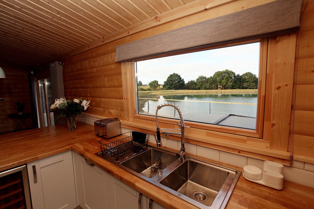 The kitchen area features a wooden countertop with a double sink beneath a large window, providing a view of the tranquil lakeside. The space is designed with light cabinetry and includes a dish drying rack. Fresh flowers add a touch of natural beauty.