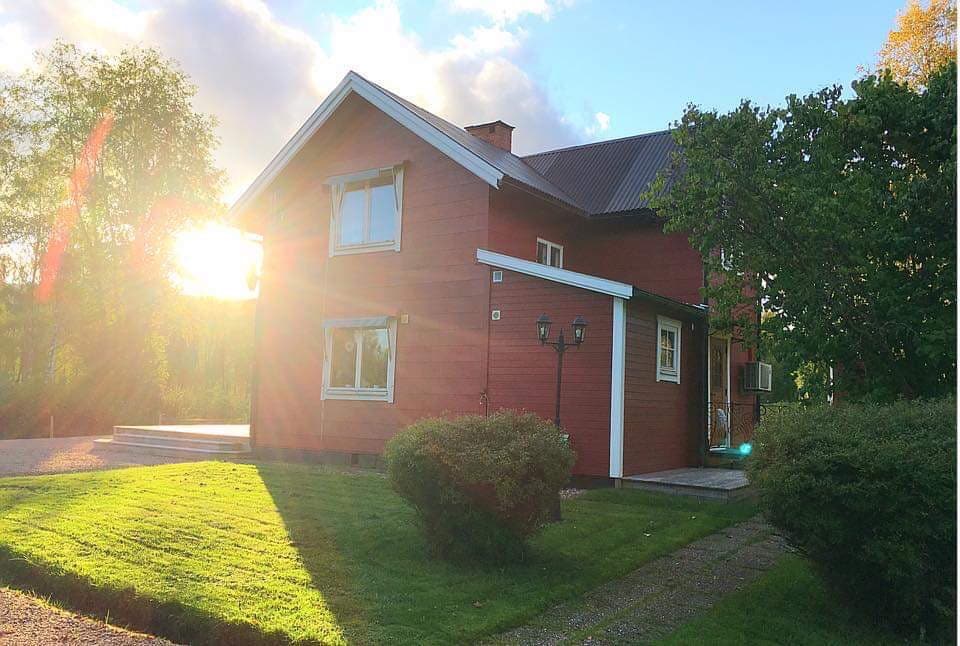 A red wooden house with a gabled roof is set against a backdrop of soft sunlight. Green grass and shrubs surround the house, while a stone pathway leads to the entrance. The structure features large windows that reflect the light.