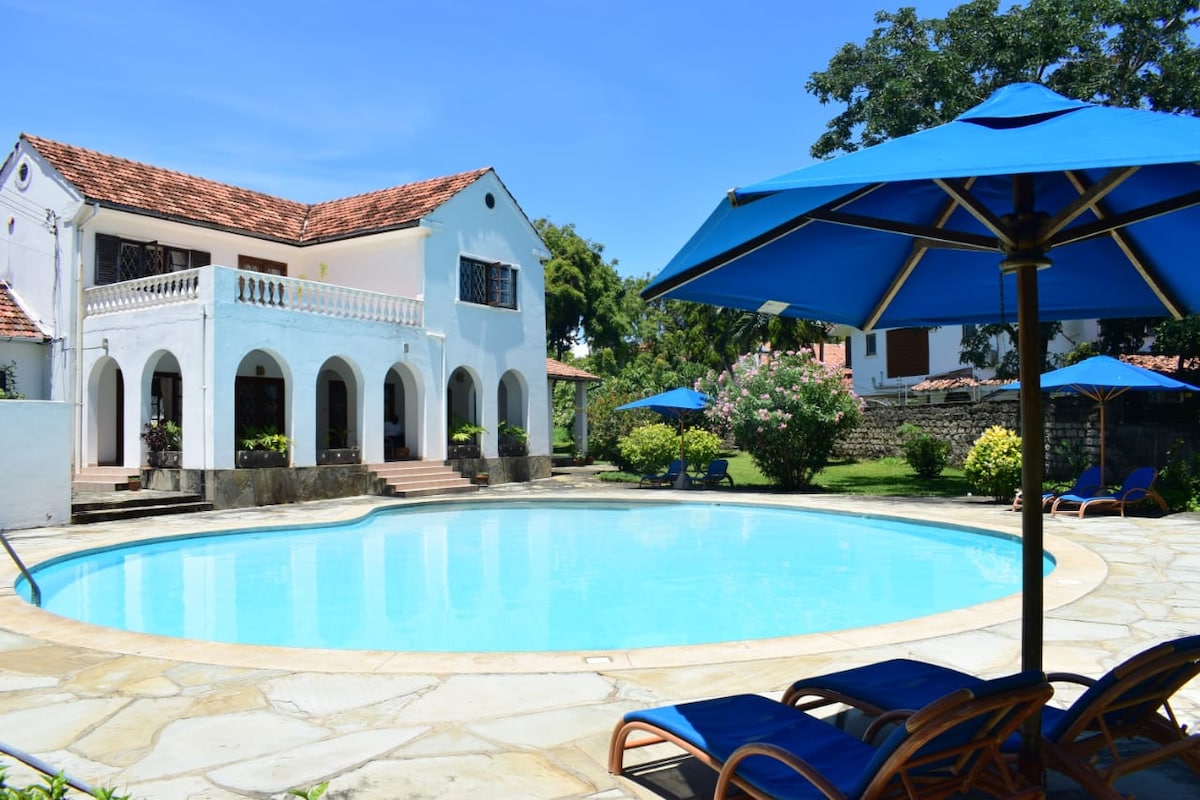 A spacious swimming pool surrounded by a stone deck is highlighted in the image. Blue umbrellas shade comfortable lounge chairs, while the white villa with a red-tiled roof and lush greenery serves as a backdrop, enhancing the property’s serene outdoor space.