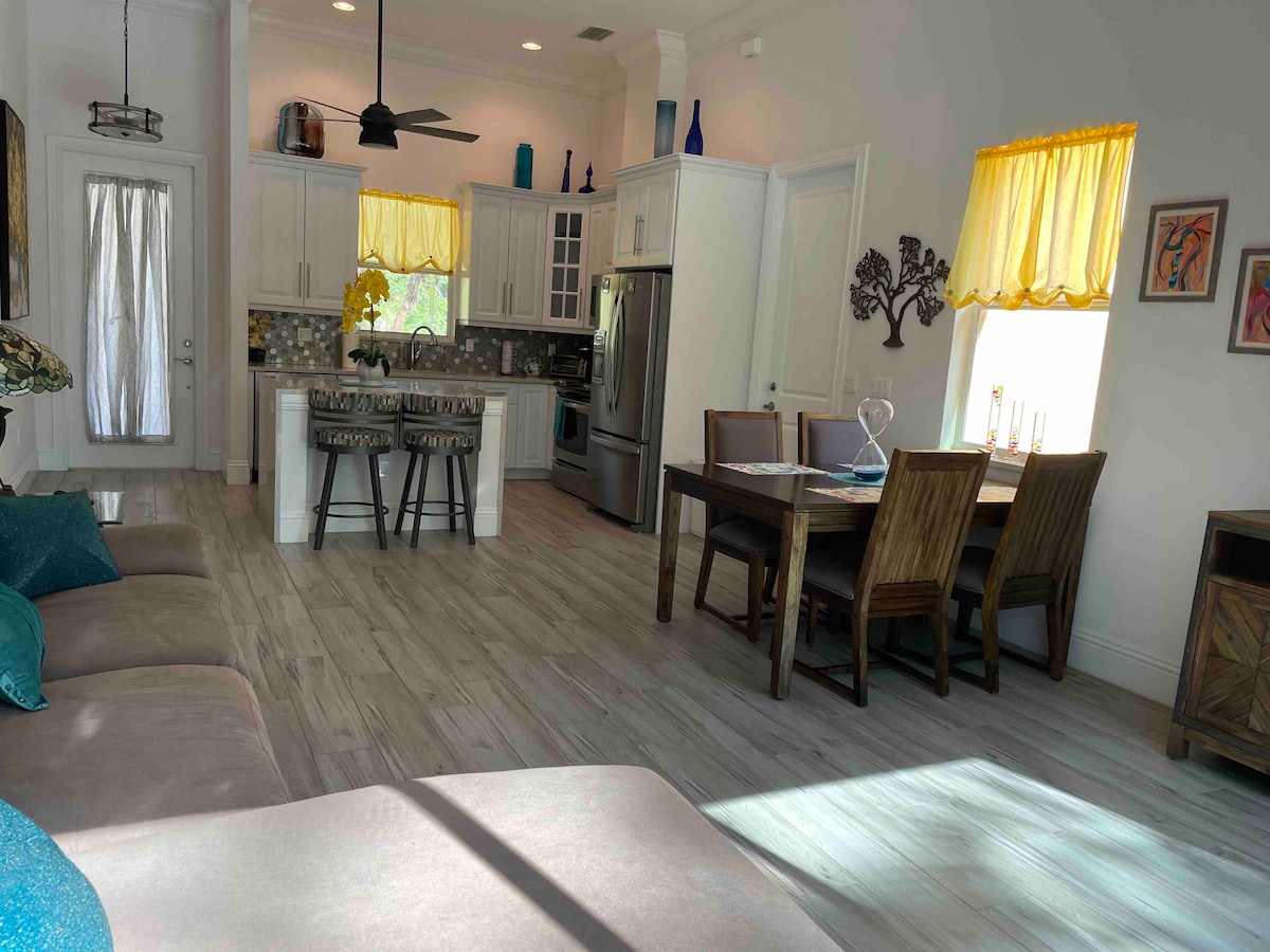 An open-concept living area features a comfortable sofa accented with blue throw pillows. A dining table accommodates multiple seats, while a well-equipped kitchen is visible in the background. Natural light fills the space through yellow curtains framing the windows.
