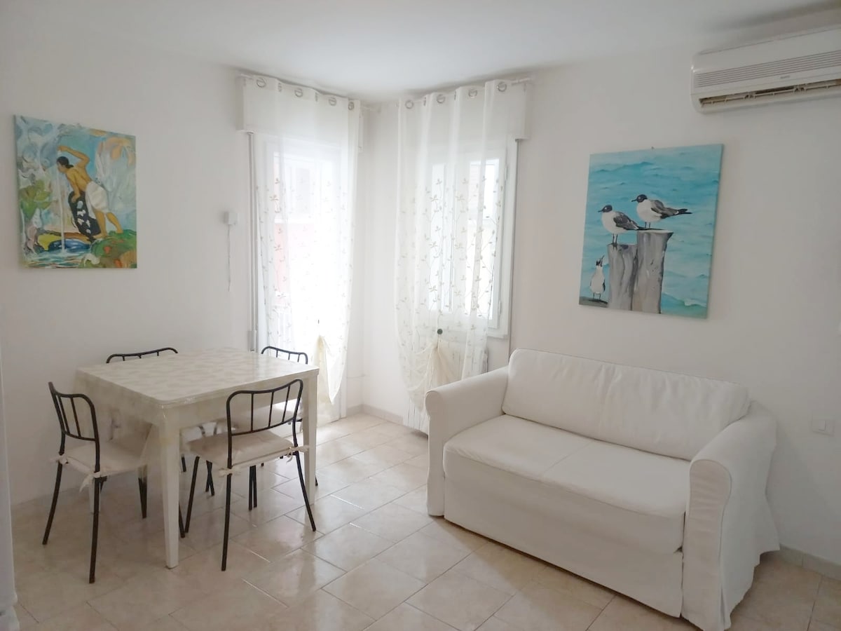 A bright living space features a white sofa and a dining table with four black chairs. Natural light filters through airy white curtains on two windows, while two pieces of colorful artwork adorn the walls, adding subtle charm to the room.