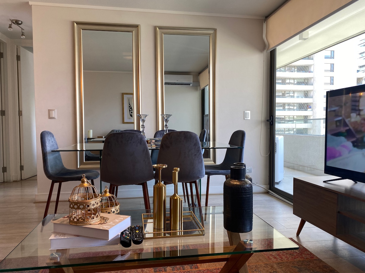 A modern dining area features a glass table surrounded by dark, upholstered chairs. Two large mirrors reflect the space, while a TV sits on a wooden console. A decorative arrangement of candles, bottles, and books adorns the coffee table, with sunlight illuminating the room from large windows.