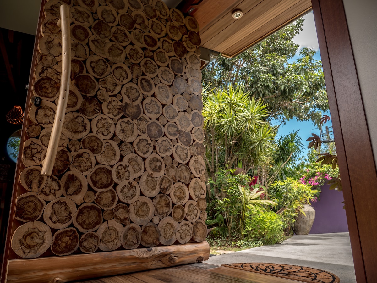 The entrance features a striking wooden wall composed of circular logs, creating a natural aesthetic. Bright greenery is visible through the open door, with hints of vibrant flowers beyond. A smooth pathway leads into the lush garden space, inviting exploration.