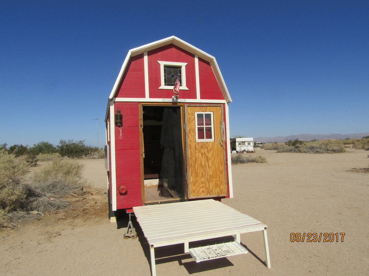 A rustic barn-style structure is depicted with a red exterior and white trim. The entrance features a wooden door that is open, revealing the interior. A small white deck leads up to the door, surrounded by desert terrain and sparse vegetation under a clear blue sky.