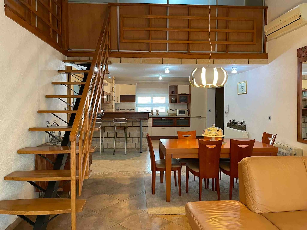 An open-plan living and dining area features a wooden staircase leading to a loft space. A rectangular dining table with six chairs is present, accompanied by a light fixture above. The kitchen is equipped with modern appliances, while a cozy seating area is visible in the foreground.