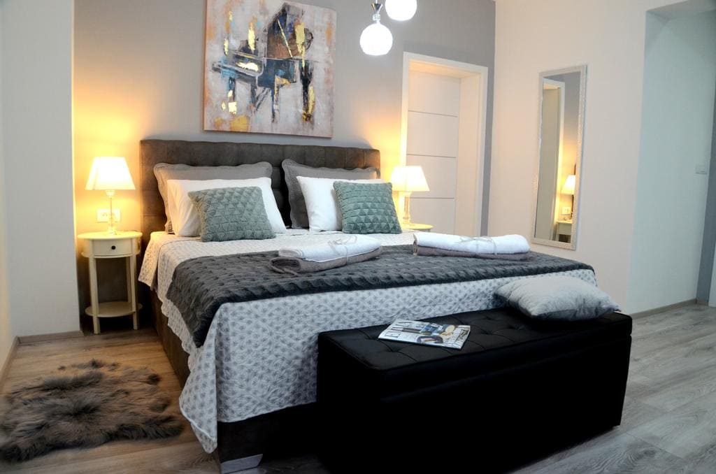 A modern bedroom features a spacious bed with crisp white linens and decorative pillows. Soft lighting is provided by bedside lamps, and a textured wall decoration adds interest. A bench sits at the foot of the bed, while a mirror reflects the room's calming design.