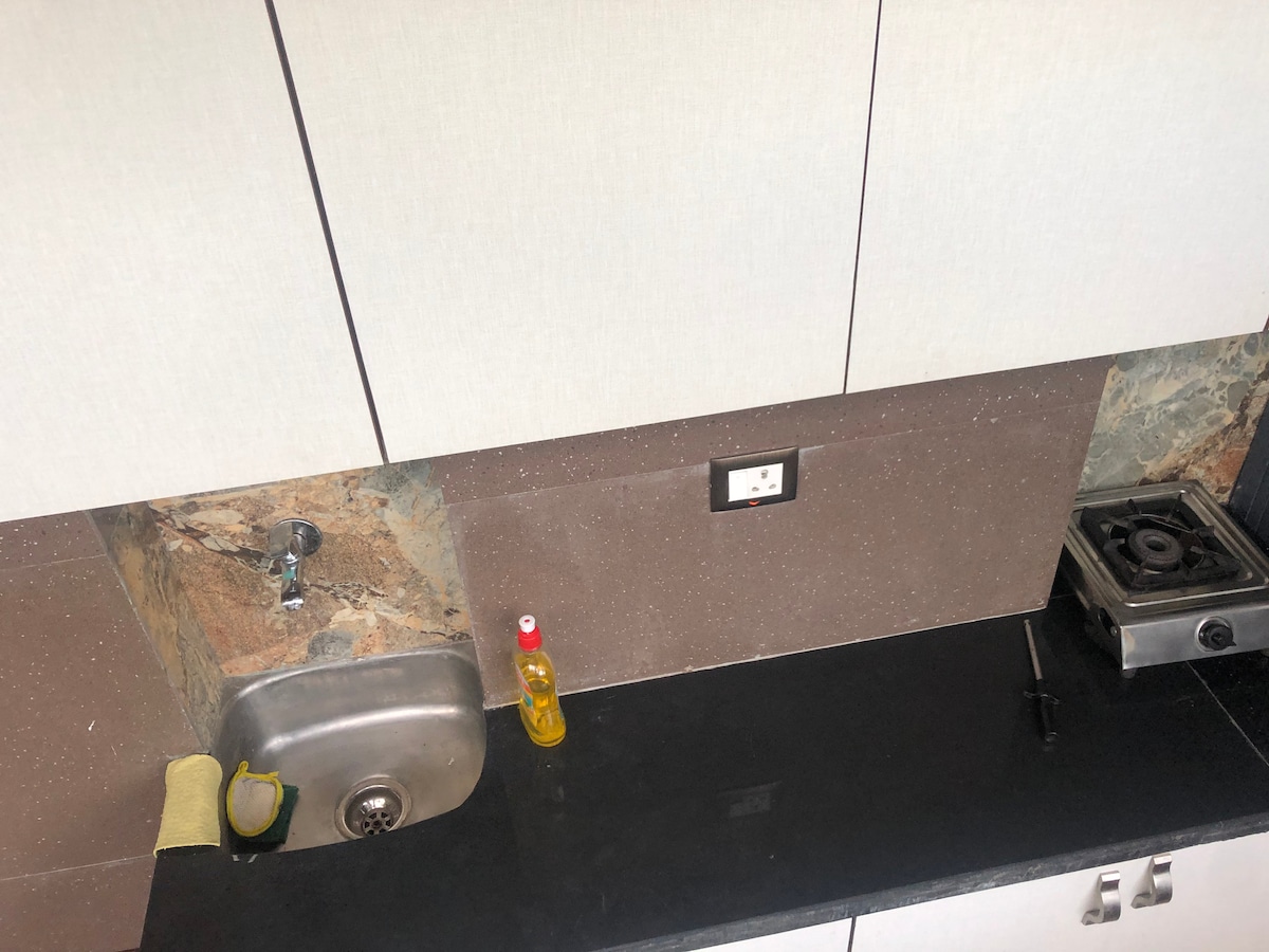 A modern kitchen setup is displayed, featuring a stainless steel sink and a marble backdrop. A compact gas stove is positioned on the right, accompanied by a small bottle of oil and a sponge on the counter, which is made of black stone.