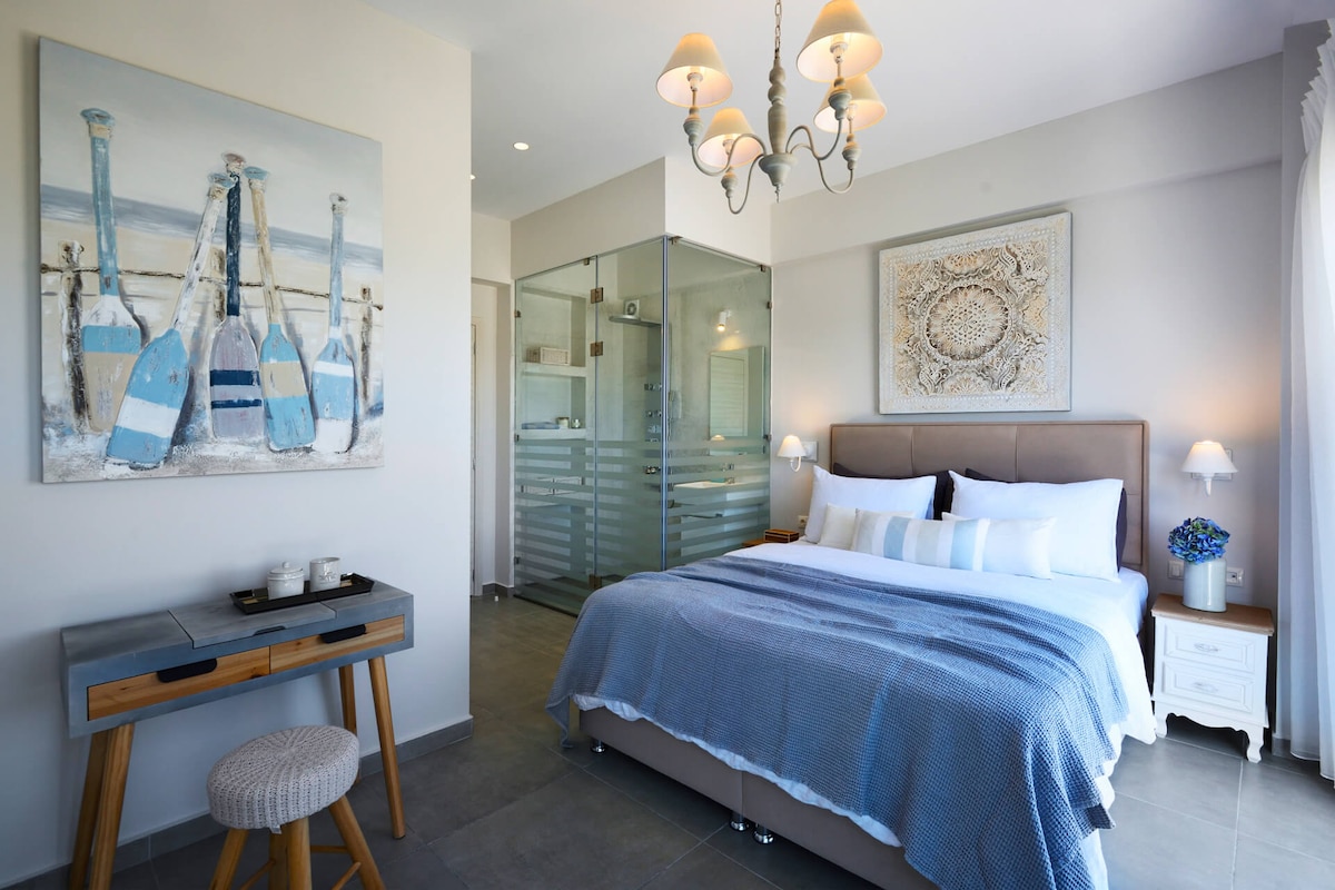 A serene bedroom features a spacious bed adorned with crisp linens and a soft blue blanket. An artistic painting of blue bottles hangs above the bed. Natural light brightens the space, highlighting the modern decor and the en-suite glass shower area.