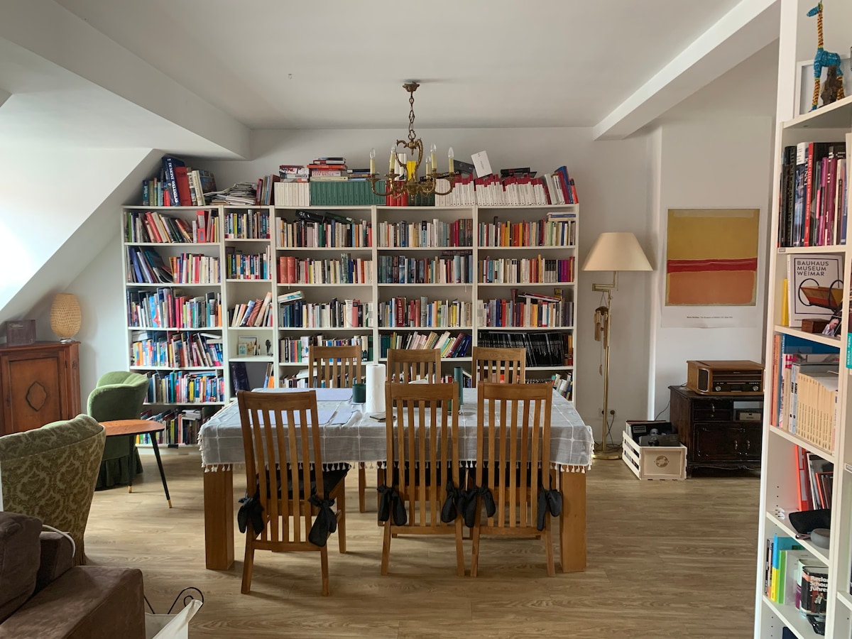 A spacious room is adorned with tall bookshelves filled with colorful books. A large wooden dining table is surrounded by six wooden chairs. A floor lamp stands in one corner, and a cozy seating area with a green chair can be seen to the left.