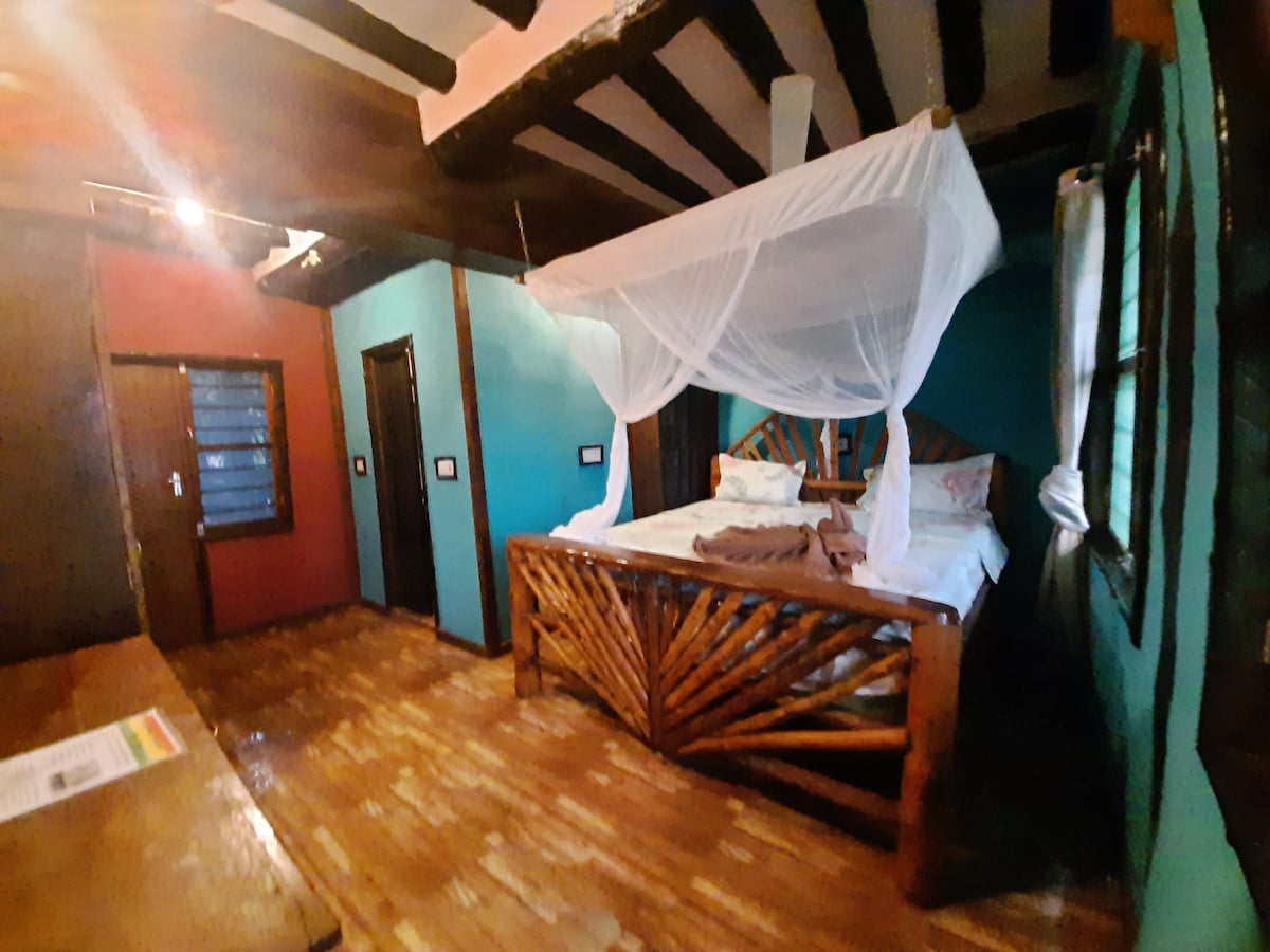 A spacious bedroom features a unique wooden bed frame with a mosquito net suspended overhead. Vibrant blue and red walls create a colorful ambiance. Natural light filters through a window, highlighting the warm wooden floor and the minimalistic furnishings, enhancing the inviting atmosphere.