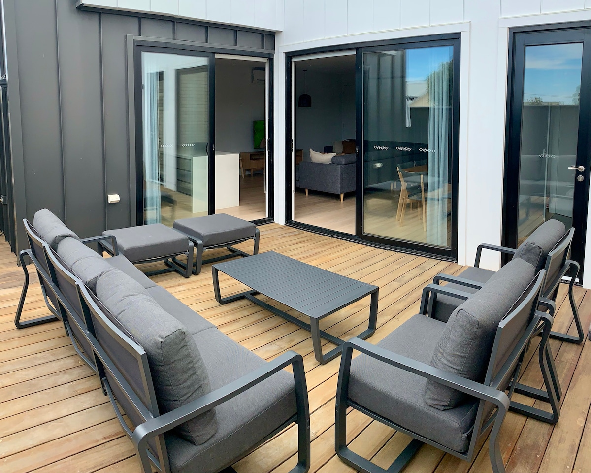 An outdoor courtyard features a modern seating arrangement consisting of two sofas, two lounge chairs, and a central coffee table, all in muted grey tones. Large glass doors allow natural light to enter from the adjoining interior space.