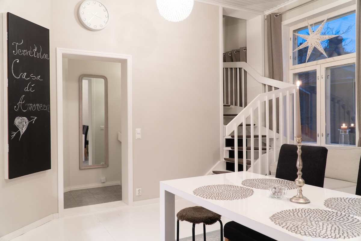 A well-lit dining area features a white table set with decorative placemats and a candleholder. A staircase is visible in the background, accompanied by a mirror reflecting natural light. A chalkboard sign adds a personal touch to the neutral-toned walls.
