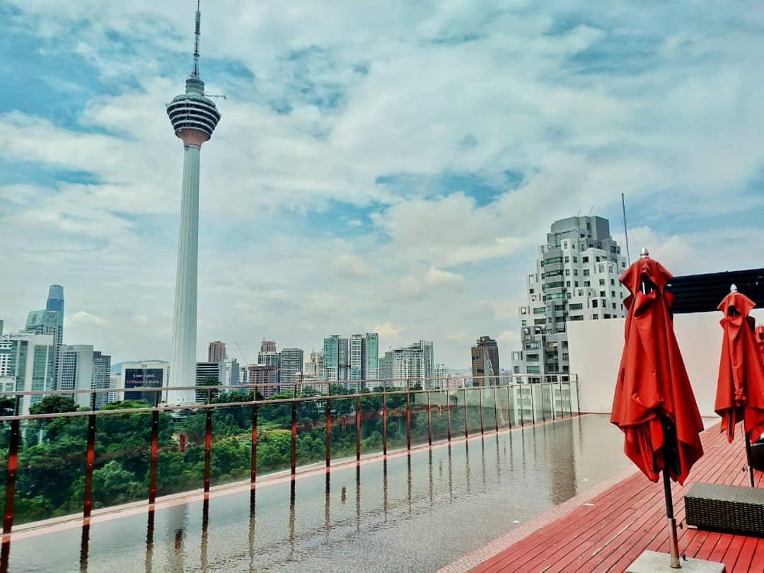 A rooftop area features a stunning view of the KL Tower amidst a city skyline. Two red umbrellas are positioned on a wooden deck, complementing the atmosphere. The surrounding landscape includes greenery and modern buildings, creating a serene escape in an urban setting.